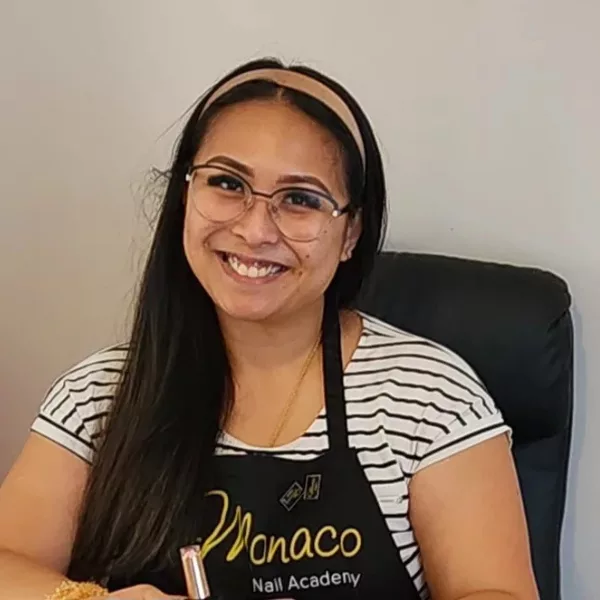 Nail Technician Pat Oyson sits at her desk with a big grin wearing a Monaco Nail Academy apron.